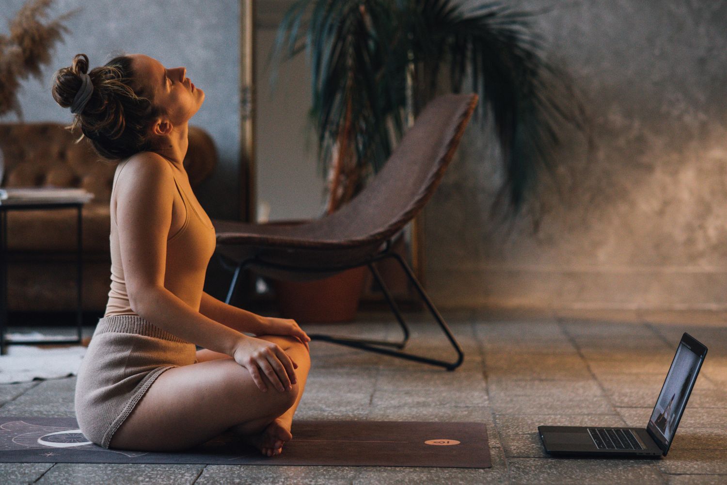 Nainen  istuu jumppamatolla ja on tekemässä pilates harjoitusta. Edessä tietokone.