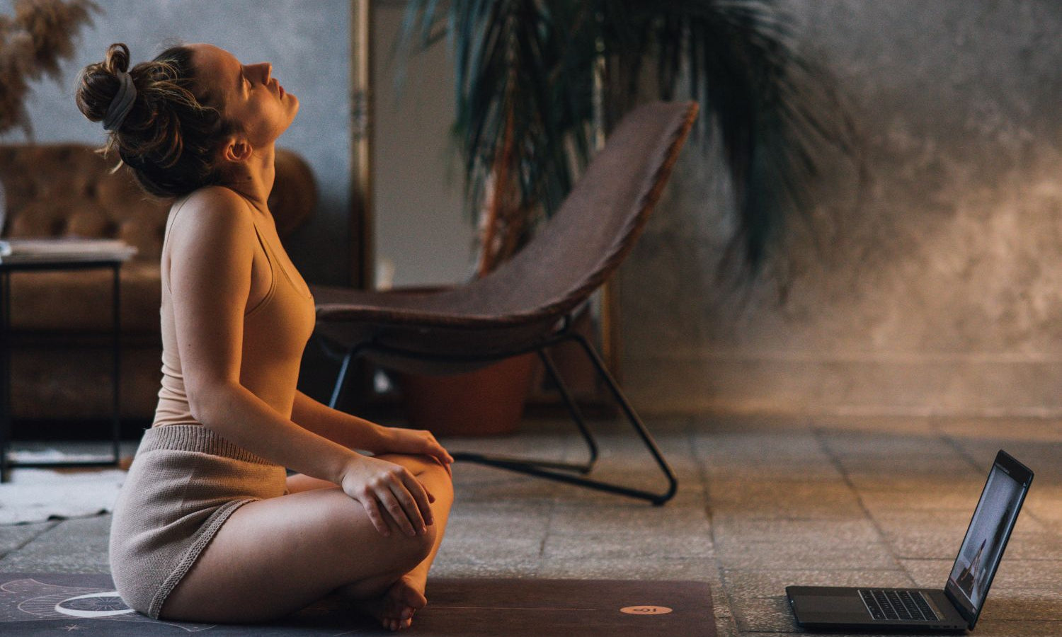 Nainen  istuu jumppamatolla ja on tekemässä pilates harjoitusta. Edessä tietokone.